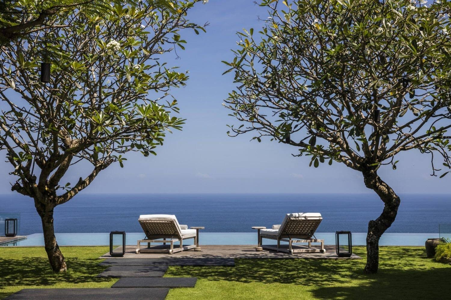 Uluwatu Estate — two loungers on the lawn between frangipani trees, the sea beyond · Uluwatu, Indonesia
